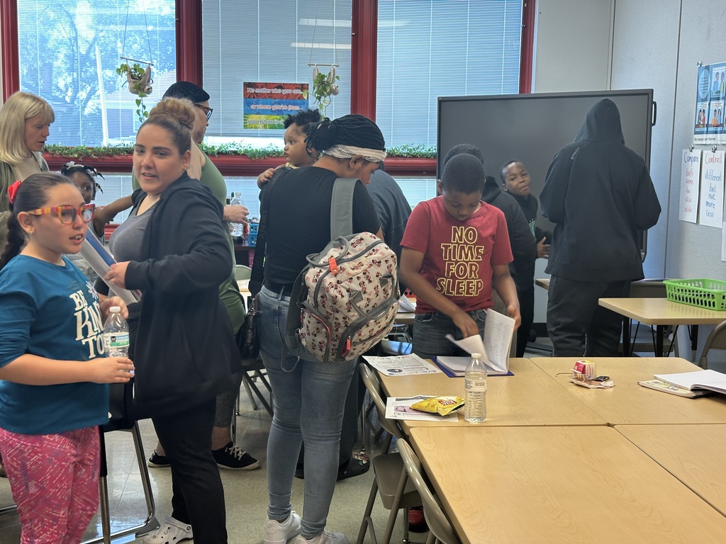 A group of students and adults interacting with one another in a classroom, with various school supplies on the tables and educational materials posted on the walls.
