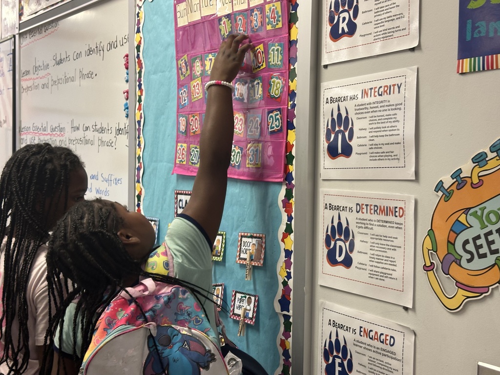A student is reaching for a number on a colorful chart in a classroom decorated with educational posters and learning objectives.