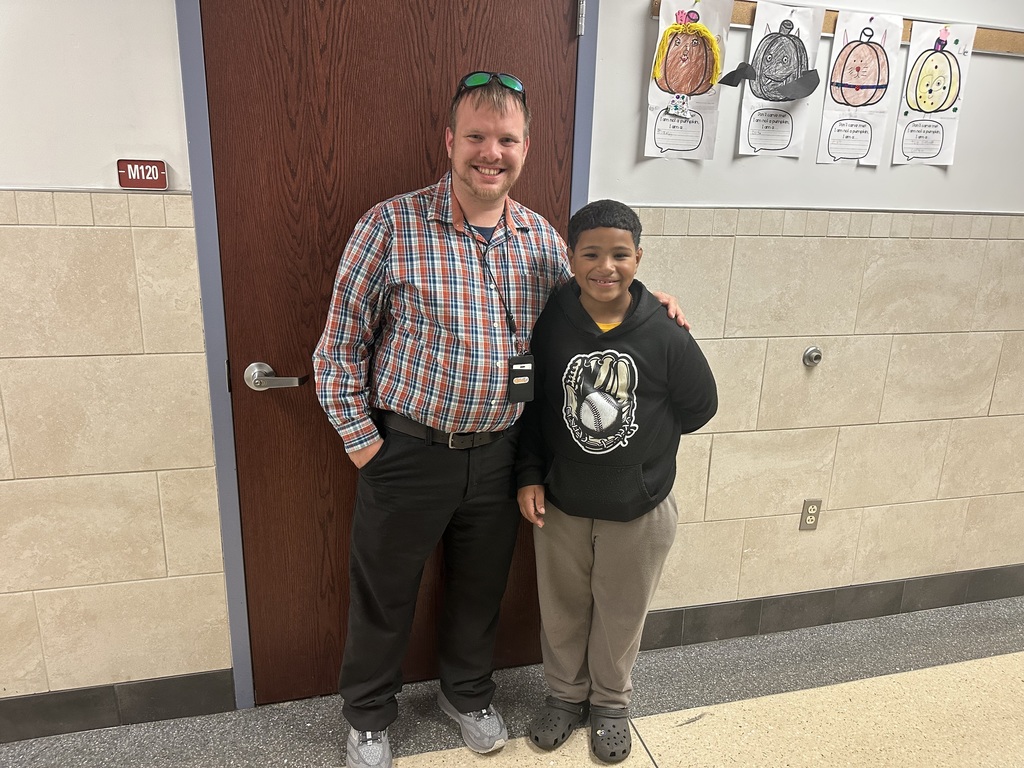 A district staff member is standing next to a student in a school hallway with artwork displayed on the wall behind them.