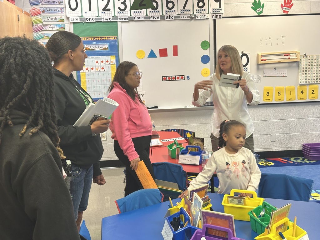 A district staff member is engaging in a conversation with a group of adults in a classroom. A student can be seen nearby at a table that has educational materials on it. 