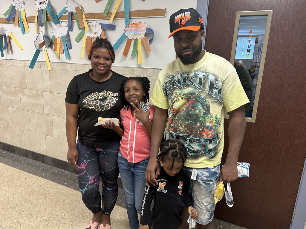 Two adults and two children standing in a school hallway in front of a wall with colorful paper crafts hanging on the wall. 