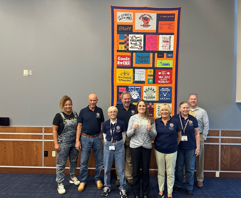 District leadership and the members of the ELKs Lodge #213 standing in front of a colorful quilt displaying various messages and schools in the School District of the City of York. One of the district staff members is holding a check in her hand.