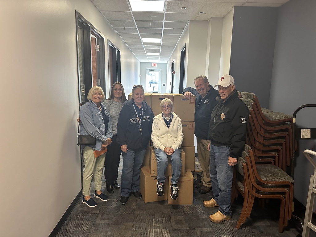District leadership and the members of the ELKs Lodge #213 standing around stacked cardboard boxes in a hallway, with stacked chairs nearby. One of the woman is sitting on one of the cardboard boxes. One of the men has his hand on one of the cardboard boxes.