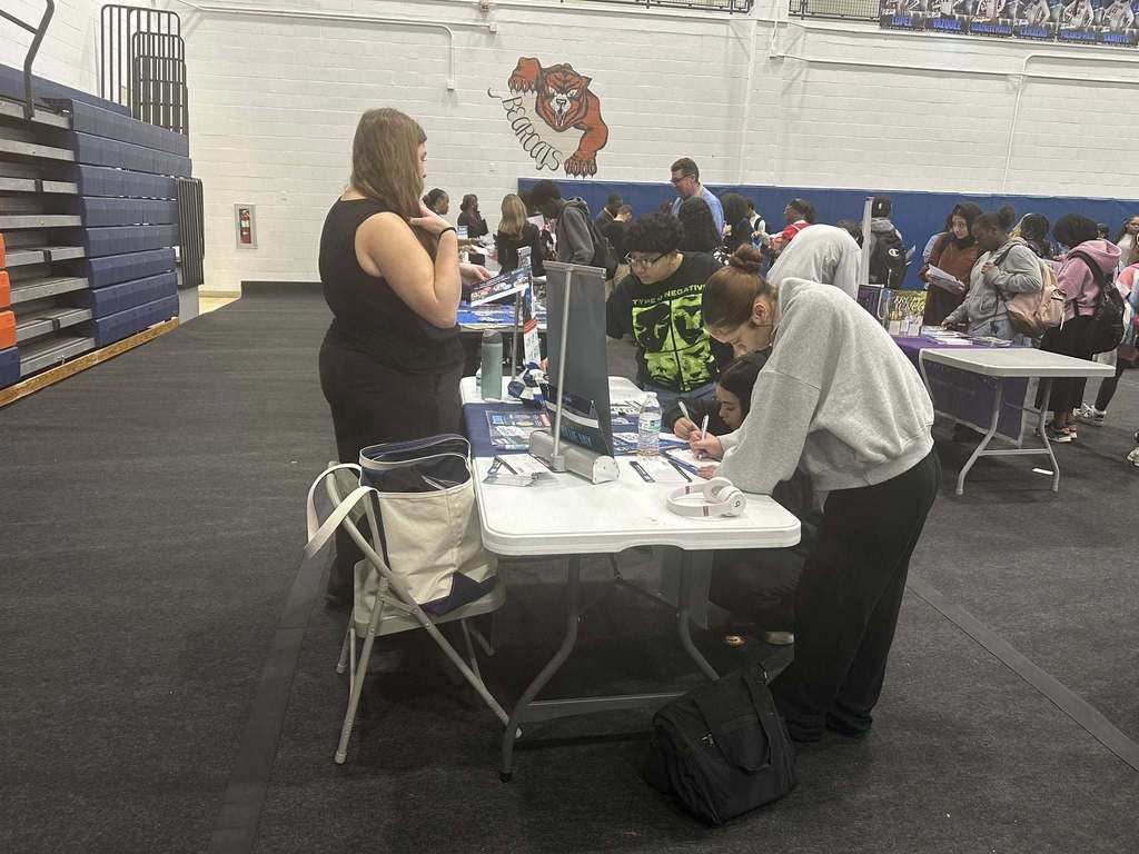 A group of students gathered around a table are engaging in a discussion with a representative at a college fair in a school gym. Two of the students are writing on a piece of paper while another student is looking at a poster. Other people can be seen in the background.