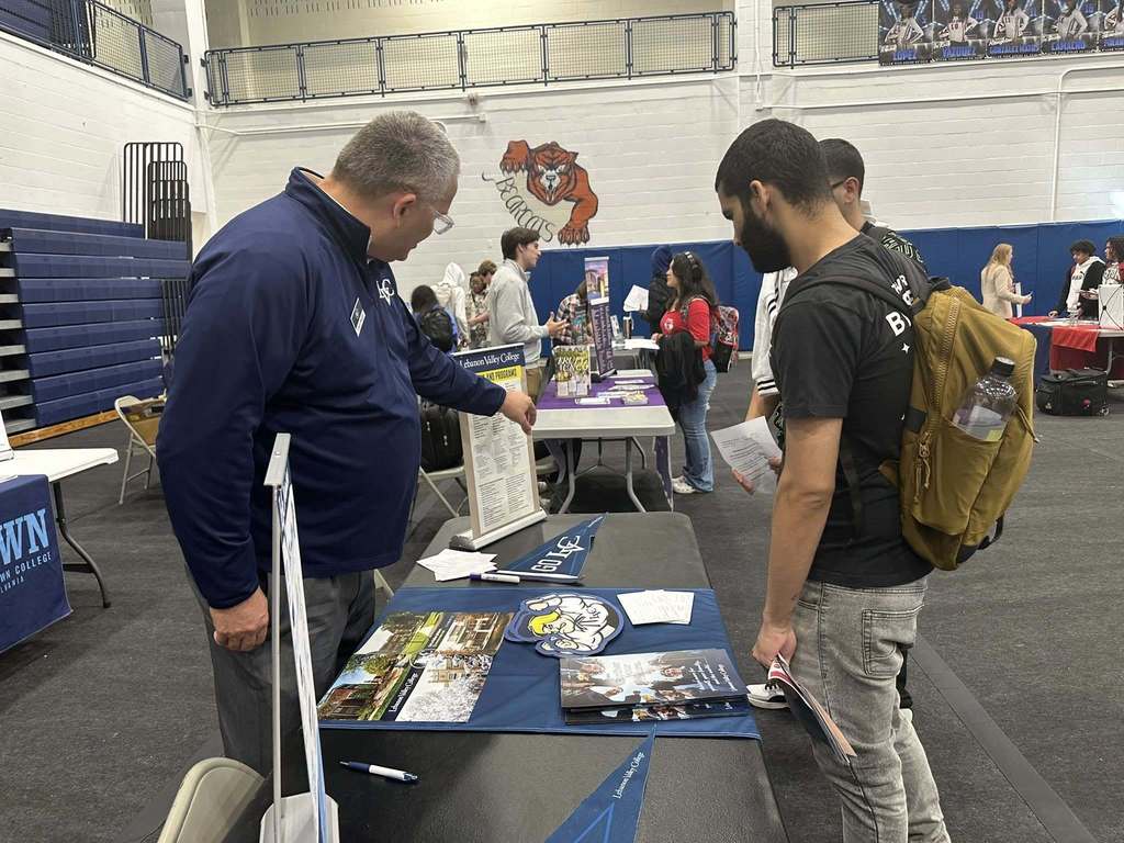 Two students and a representative are engaging in a discussion at a college fair in a school gym. Other people can be seen in the background.