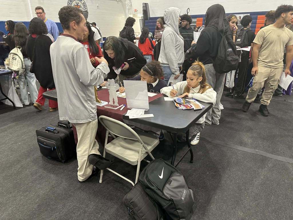 A group of students gathered around a table are engaging in a discussion with a representative at a college fair in a school gym. The students are writing on a piece of paper while other people can be seen in the background.