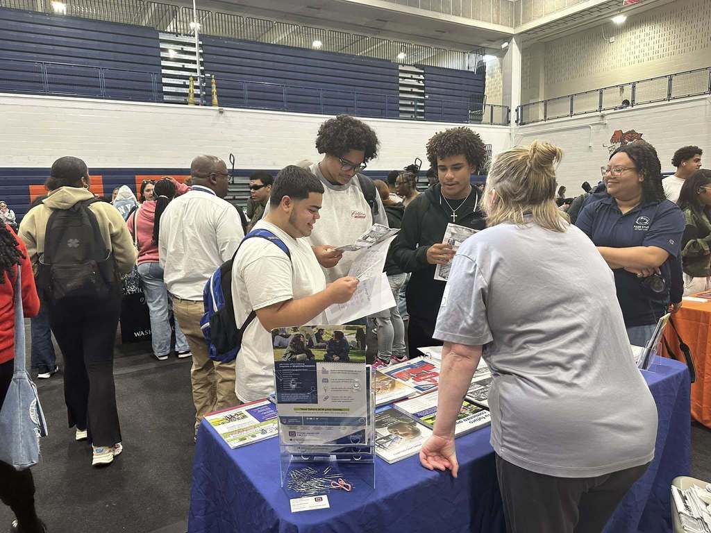 A group of students are engaging with a representative at a college fair in a school gym. Other people can be seen in the background.