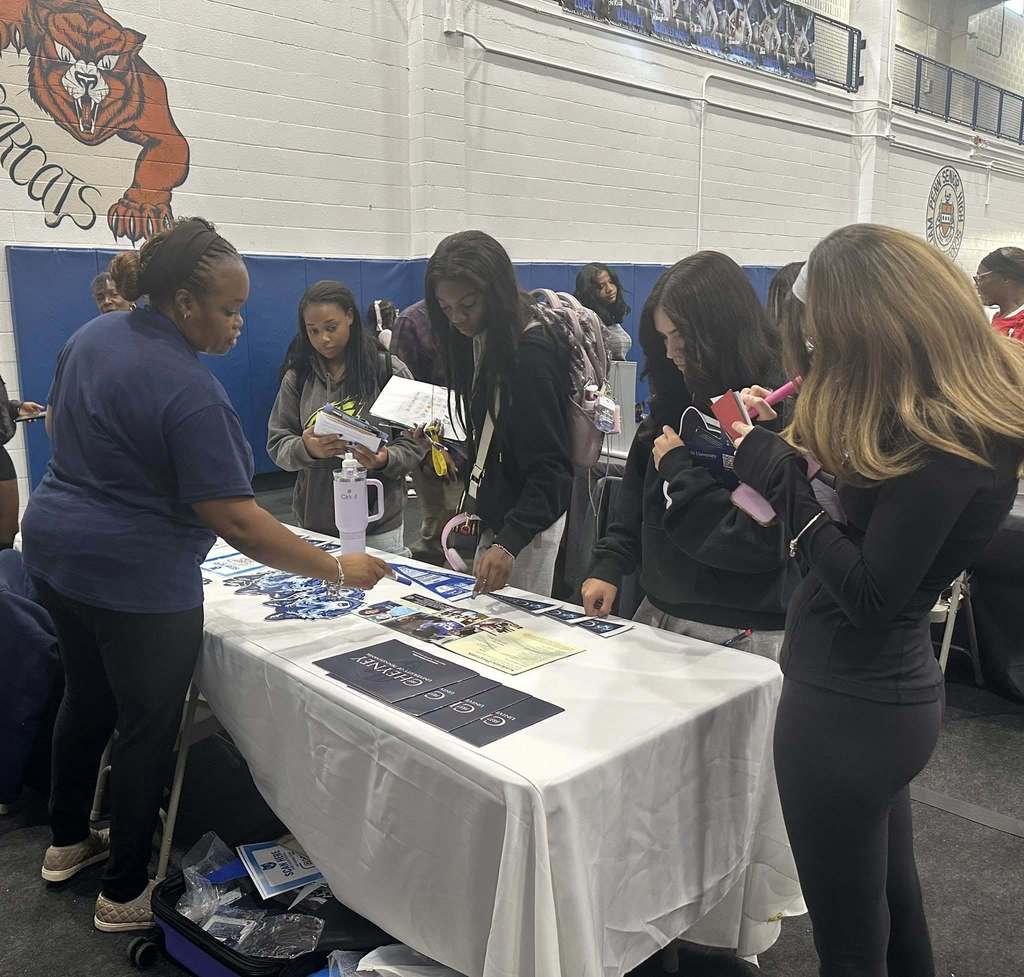 A group of students are engaging with a representative at a college fair in a school gym, exploring informational materials on a table. Other people can be seen in the background.