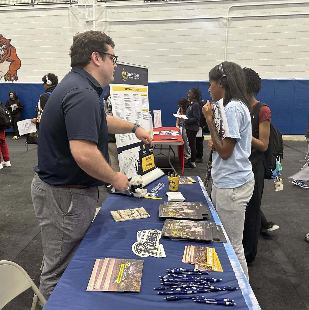 A student and a representative are engaging in a discussion at a college fair in a school gym. Other people can be seen in the background.
