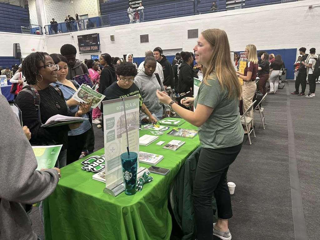 A group of students are interacting with a representative at a college fair in a school gym. Other people can be seen in the background.
