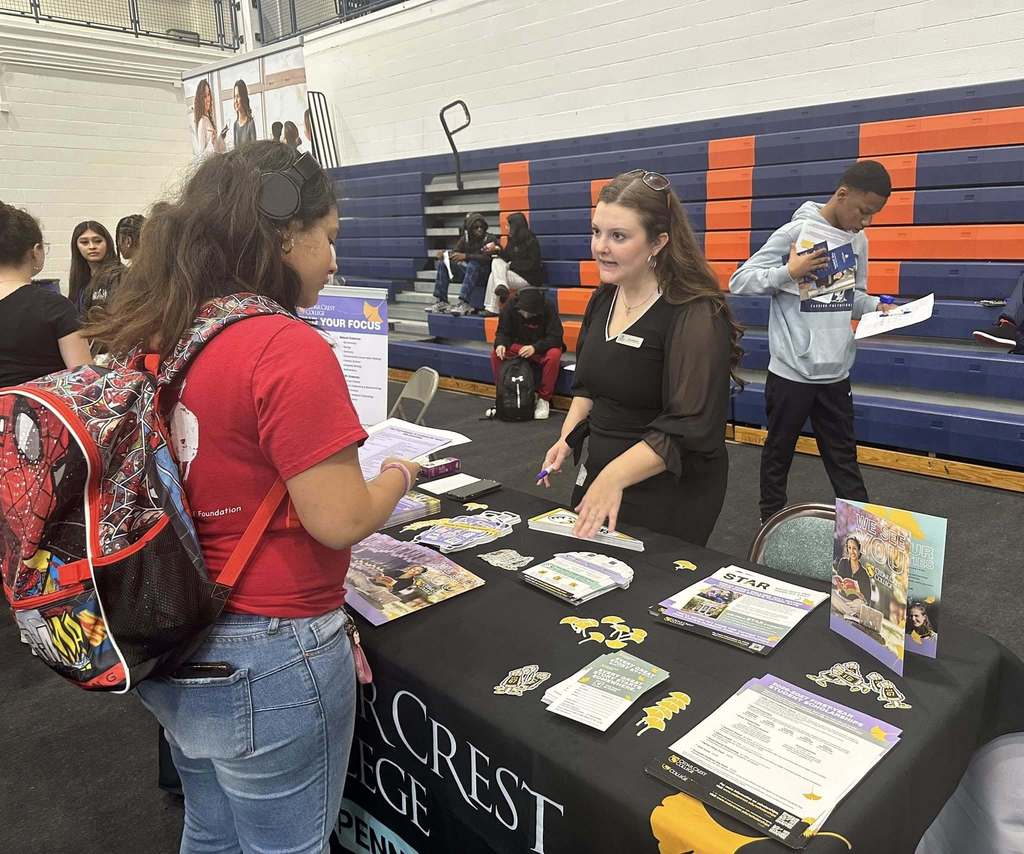 A student and a representative are engaging in a discussion at a college fair in a school gym. Other people can be seen in the background.