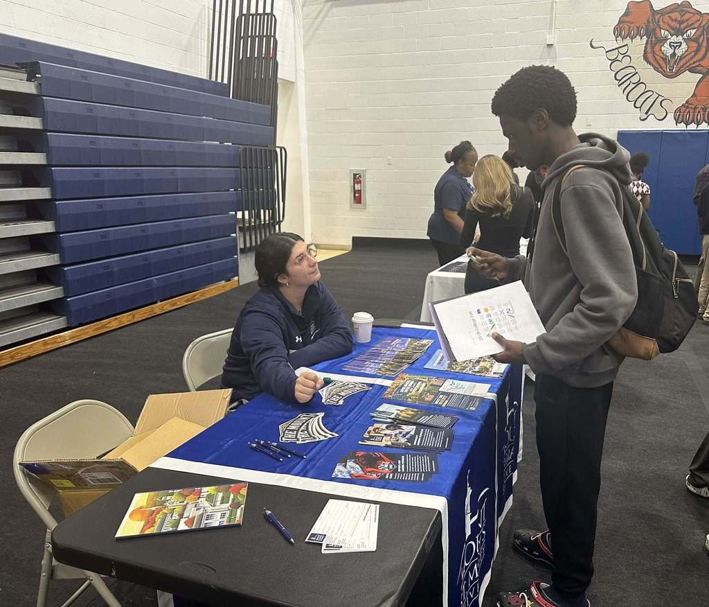 A student and a representative are engaging in a discussion at a college fair in a school gym. Other people can be seen in the background.