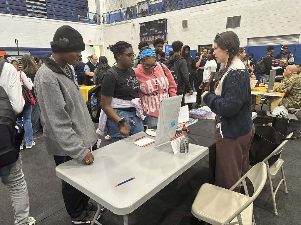 A group of students and a representative are engaging in a discussion at a college fair in a school gym. Other people can be seen in the background.
