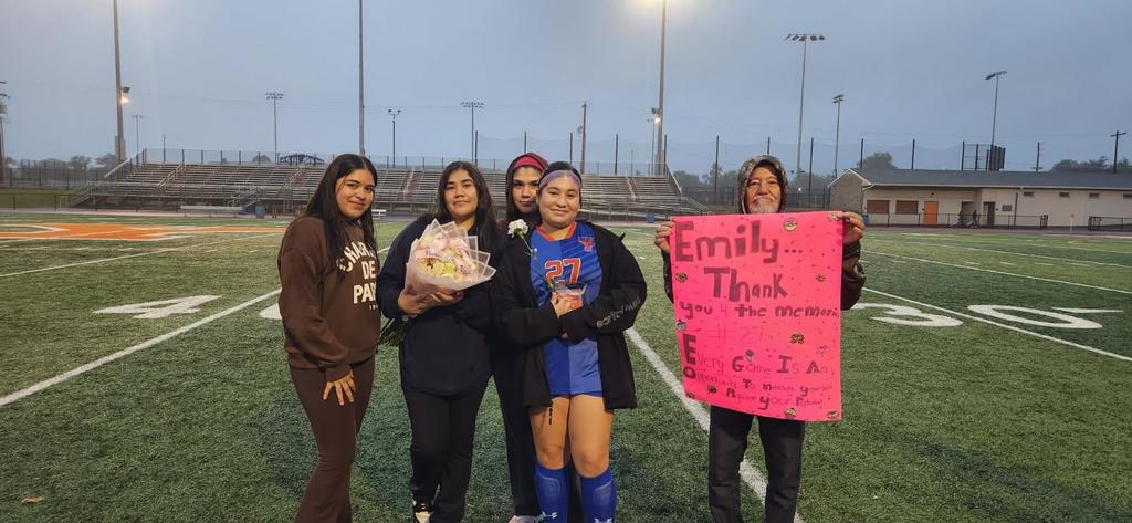 A group of five individuals are standing together outside on a soccer field, with the one person holding a trophy and a flower and another person holding a bouquet of flowers, celebrating Girls Soccer Senior Night.