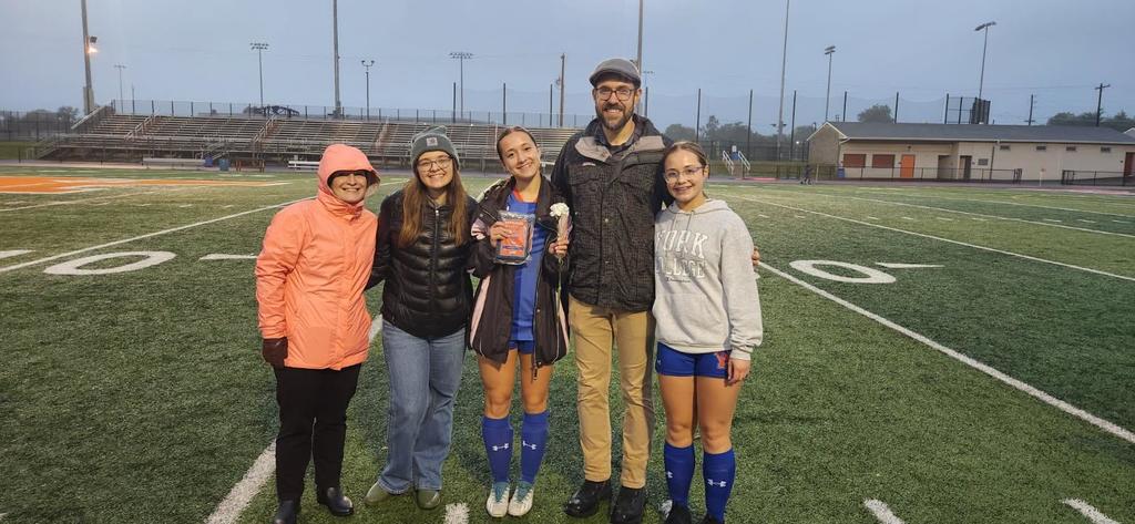 A group of five individuals are standing together outside on a soccer field, with one person holding a trophy and a flower, celebrating Girls Soccer Senior Night.
