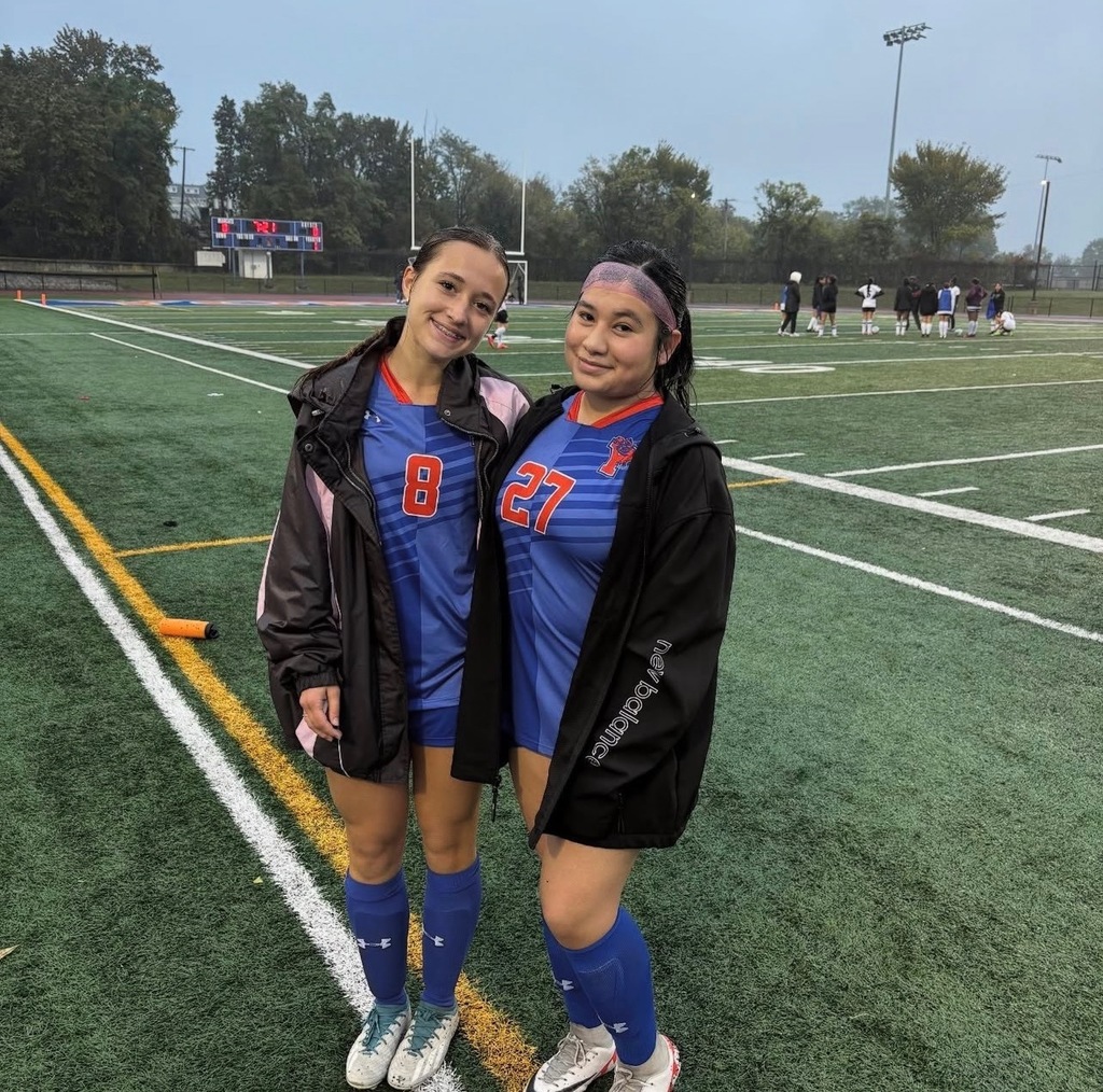Two York High Girls Soccer players are standing outside on a soccer field in blue team jerseys, one is wearing a black jacket over her uniform. Other people and a scoreboard can be seen in the background.