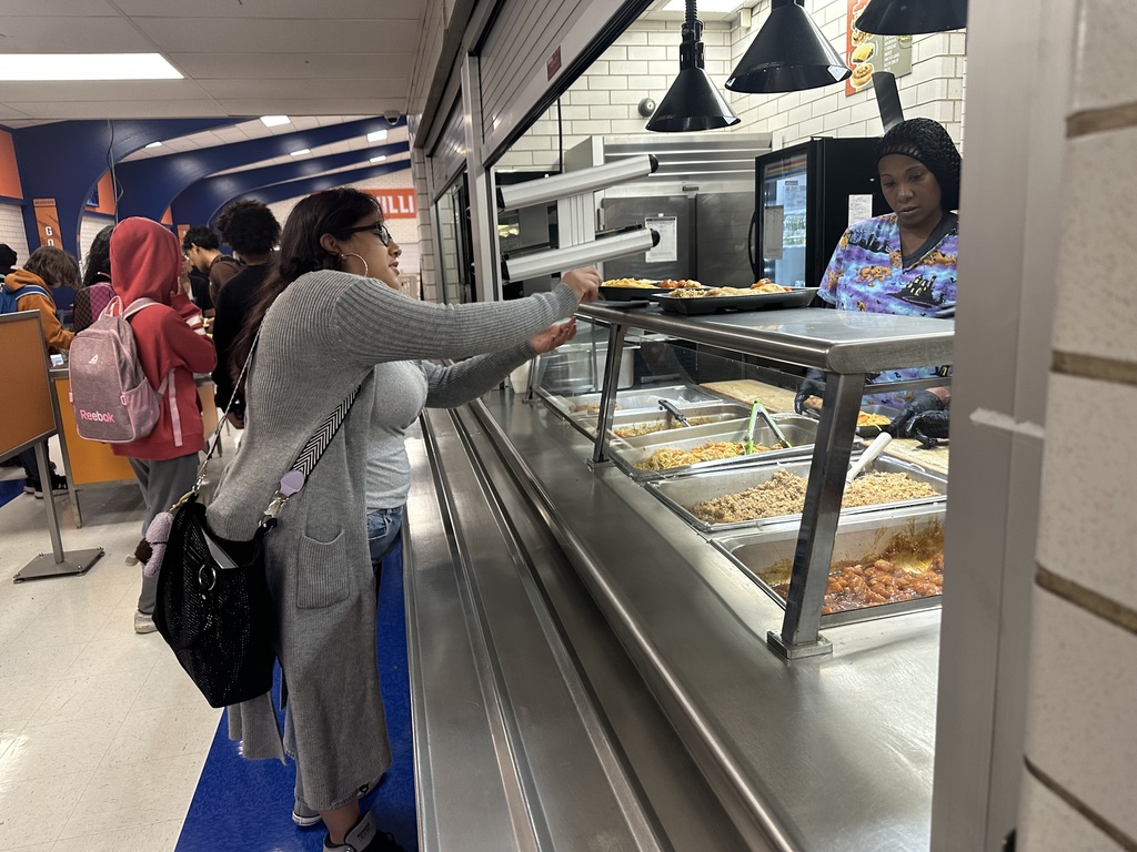 A student is reaching for a lunch tray in a school cafeteria. A district staff member wearing a hair net can be seen behind a counter. Other people are visible in the background.