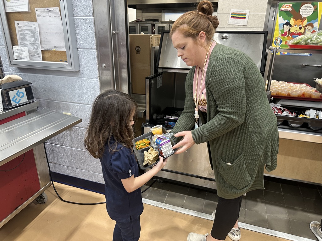 A district staff member is handing a lunch tray that has vegetables and chips on it to a student in a school cafeteria. 
