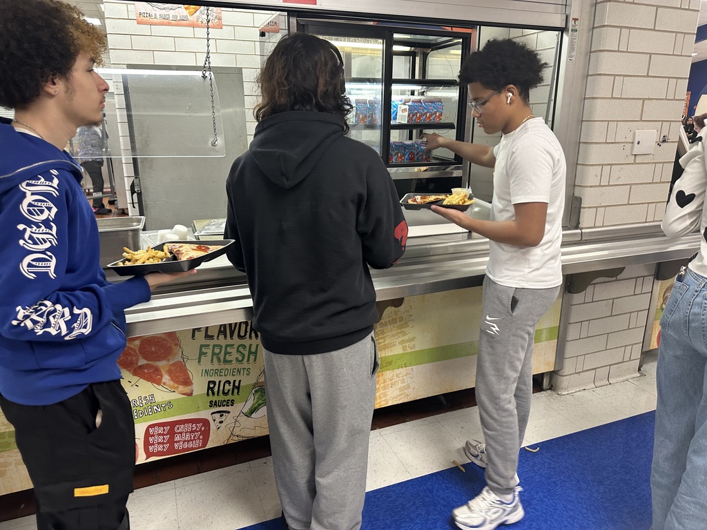 Three students are in a cafeteria line in a school cafeteria. One of the students is reaching for a milk carton from a mini fridge. 