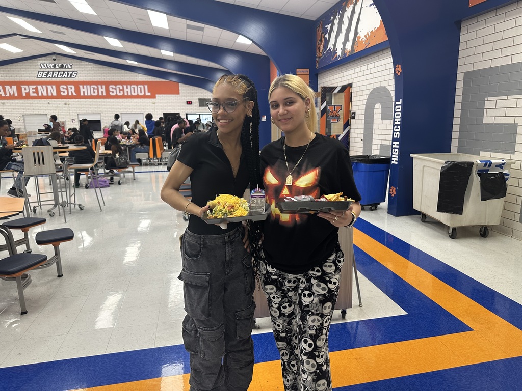 Two students are holding lunch trays in a school cafeteria, surrounded by other people. 