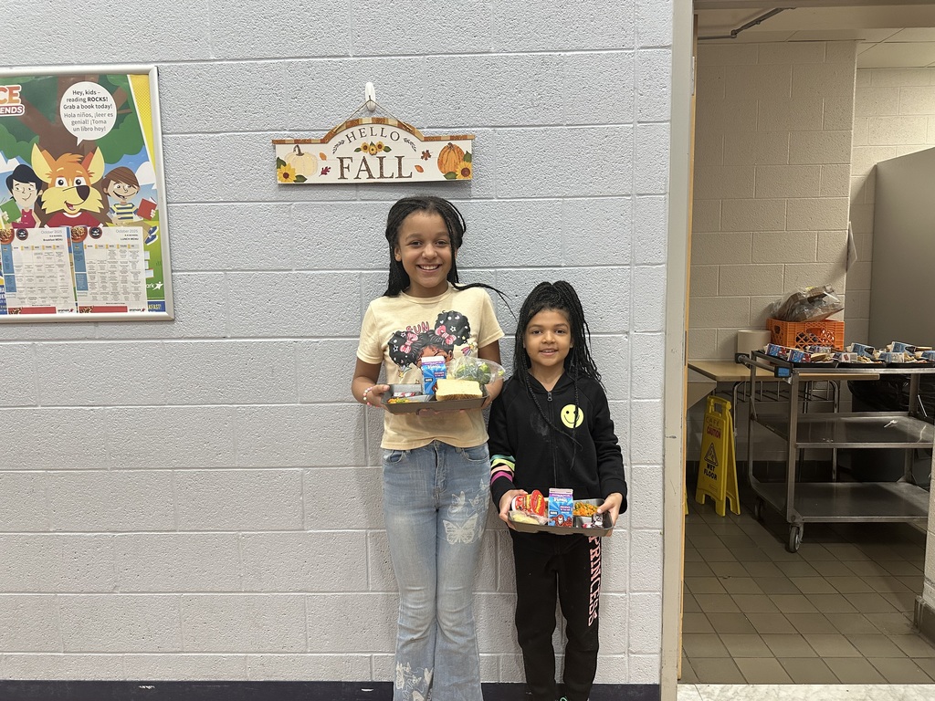 Two students holding lunch trays in a school cafeteria. A sign above them reads "Hello Fall." 