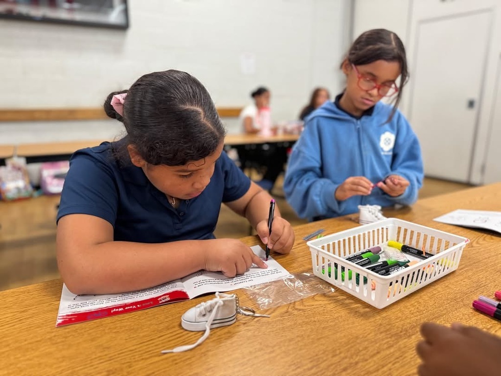 Two students are siting at a table in a school cafeteria engaging in an activity. One of the students is using a marker to draw in a booklet, while the other student is examining a small object. Other people can be seen in the background sitting at another table.