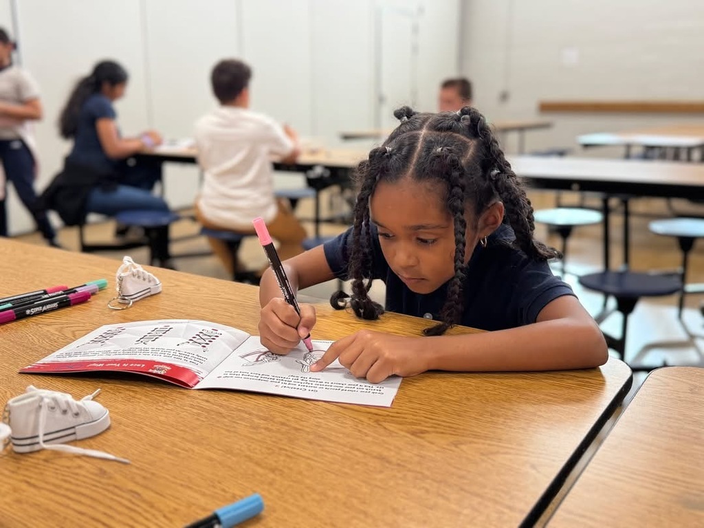 A student is sitting at a table in a school cafeteria, using a marker to trace a design in a booklet. Other people can be seen in the background sitting at another table.