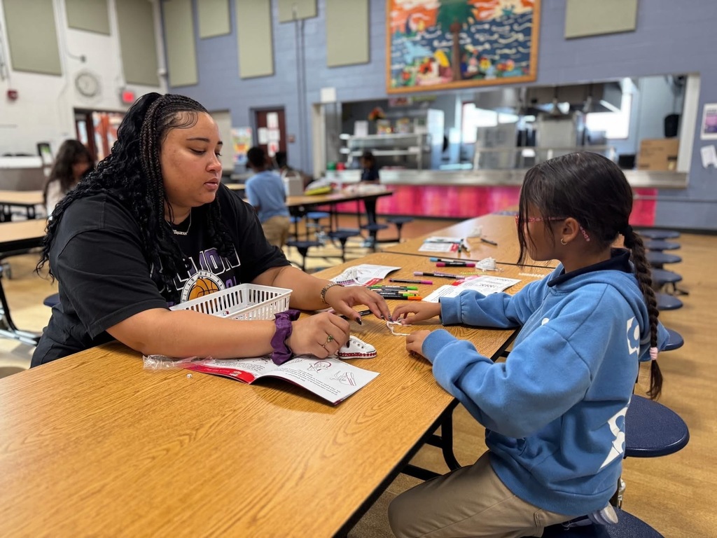 An instructor and a student are sitting at a table in a school cafeteria engaging in an activity with a booklet and markers. The instructor is helping the student with the activity. Other people can be seen in the background sitting at other tables.