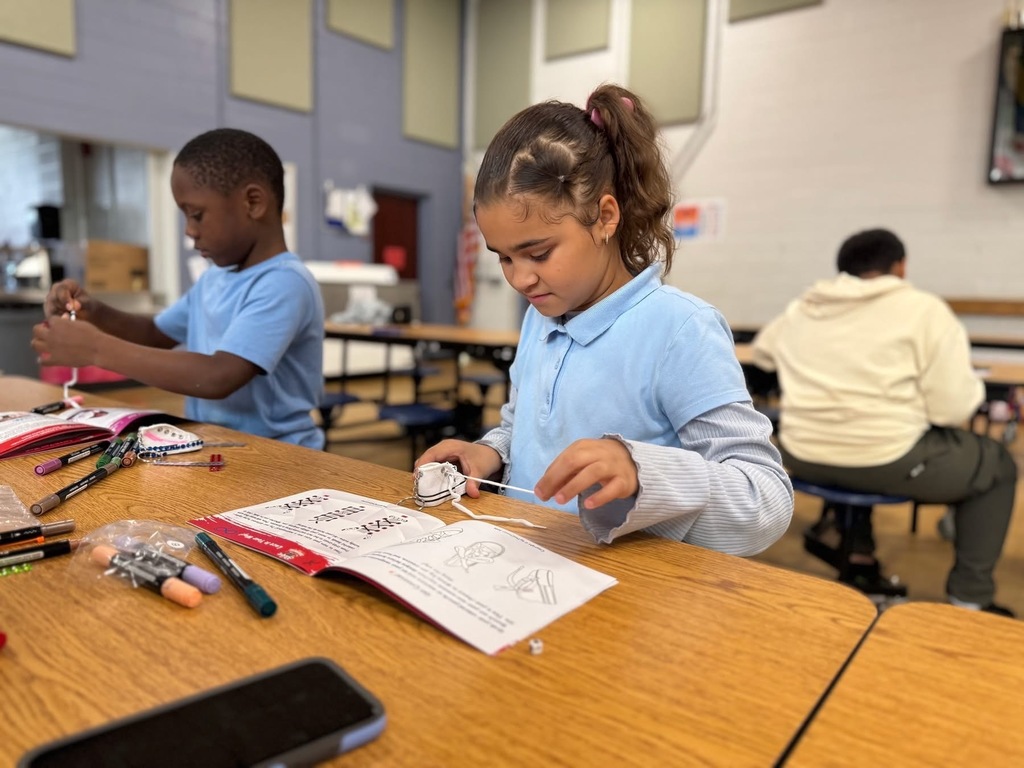 Two students are siting at a table in a school cafeteria engaging in an activity. Booklets and markets can be seen on the table nearby. Other people can be seen in the background sitting at another table.