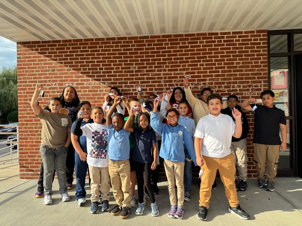 A group photo of students and one adult standing in front of a brick wall. Some of the students are holding up small objects.