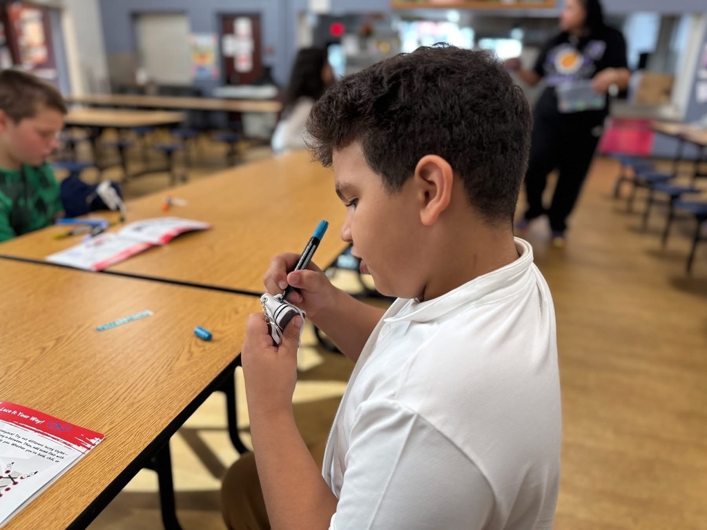 A student is sitting a table in a school cafeteria using a marker to color on a small sneaker. Other people can be seen in the background nearby.