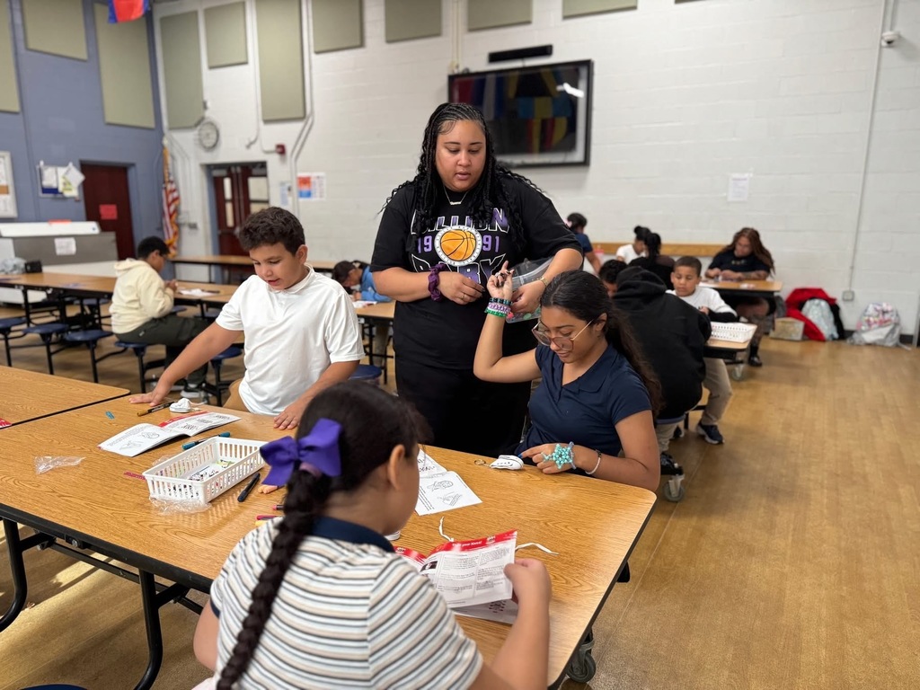 An instructor is helping students who are sitting at a table in a school cafeteria with an activity. Other people can be seen in the background sitting at other tables.