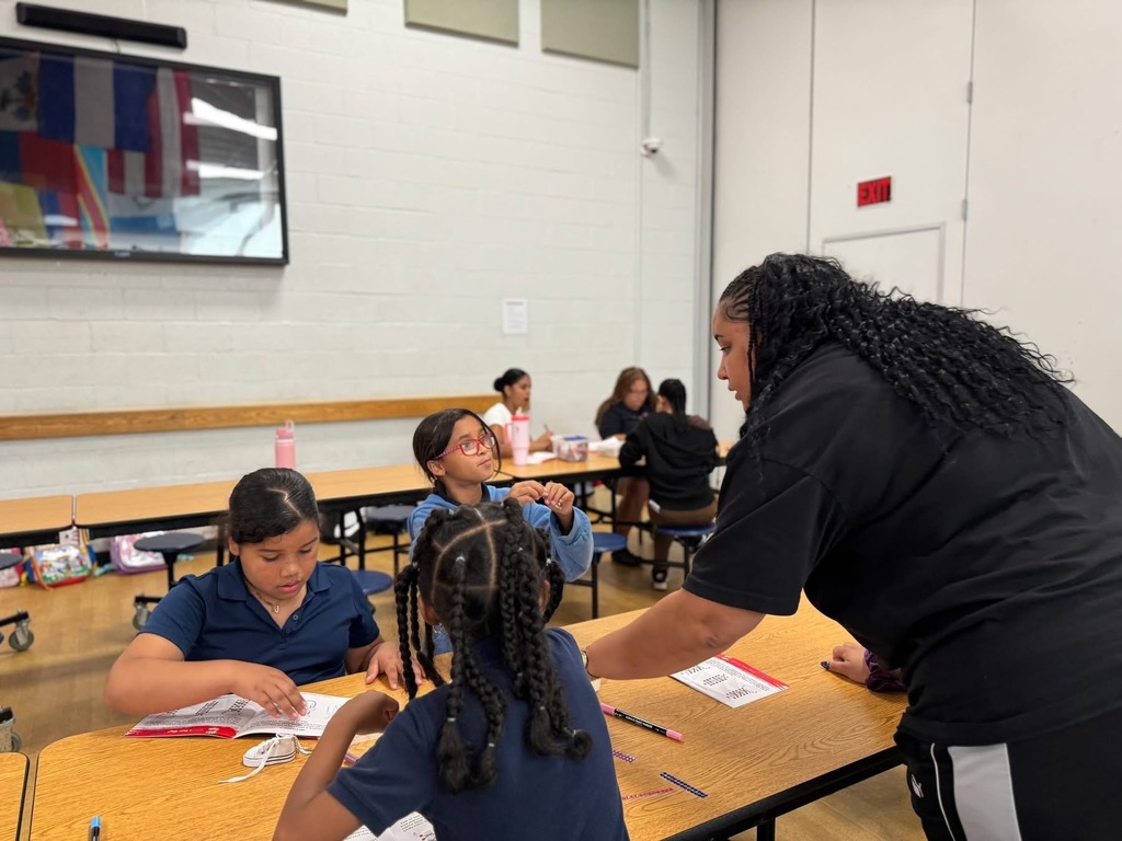 An instructor assisting a group of students at a table in a school cafeteria. Other people can be seen in the background sitting at another table.