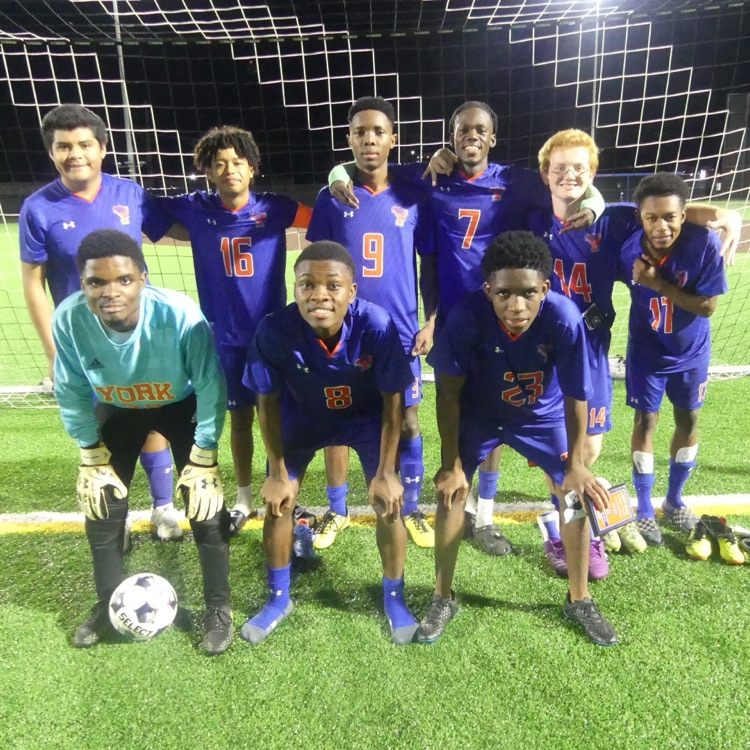 Eight York High Boys Soccer team members standing outside in front of a soccer net.