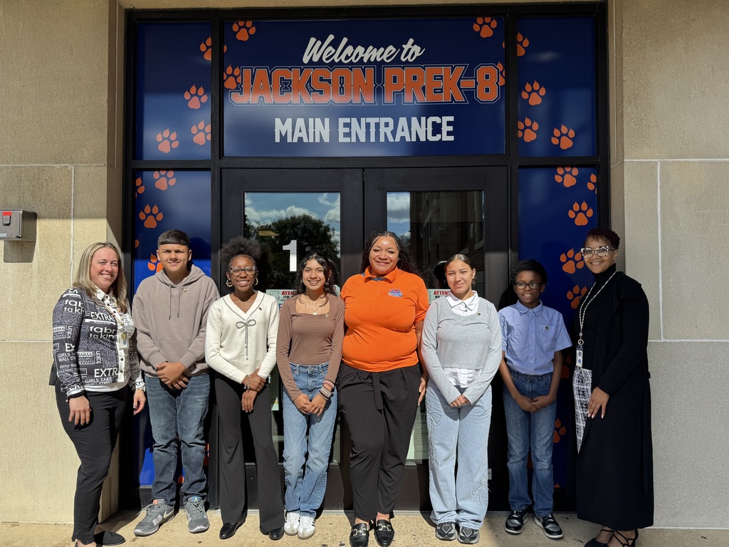 District leadership and a group of students standing under a sign that reads "Welcome to JACKSON PREK-8 MAIN ENTRANCE". 