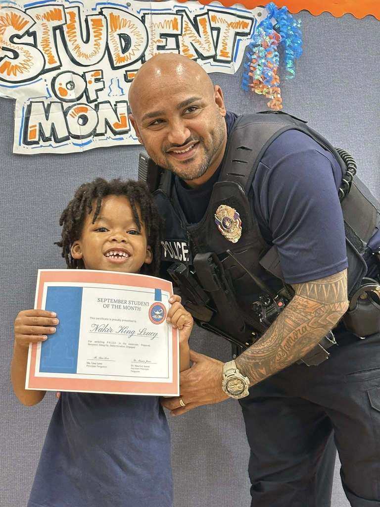 A student and a school police officer are smiling in front of a "Student of the Month" sign. The student is holding a certificate at Ferguson PreK-8's September Student of the Month Celebration.