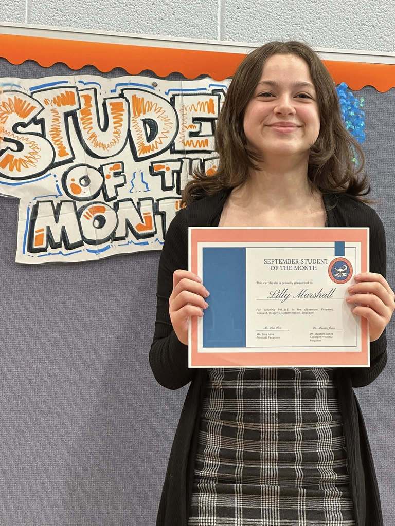 A student is standing in front of a "Student of the Month" sign, holding a certificate at Ferguson PreK-8's September Student of the Month Celebration.