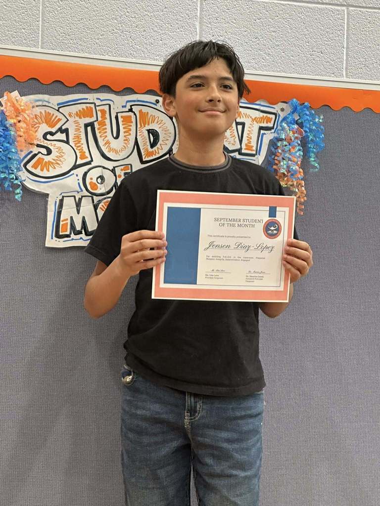 A student is standing in front of a "Student of the Month" sign, holding a certificate at Ferguson PreK-8's September Student of the Month Celebration.