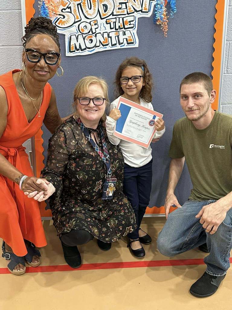 Ferguson staff, a student and an adult are smiling in front of a "Student of the Month" sign. The student is holding a certificate at Ferguson PreK-8's September Student of the Month Celebration.