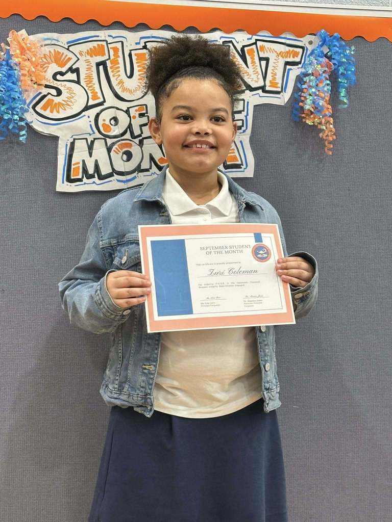 A student is standing in front of a "Student of the Month" sign, holding a certificate at Ferguson PreK-8's September Student of the Month Celebration.