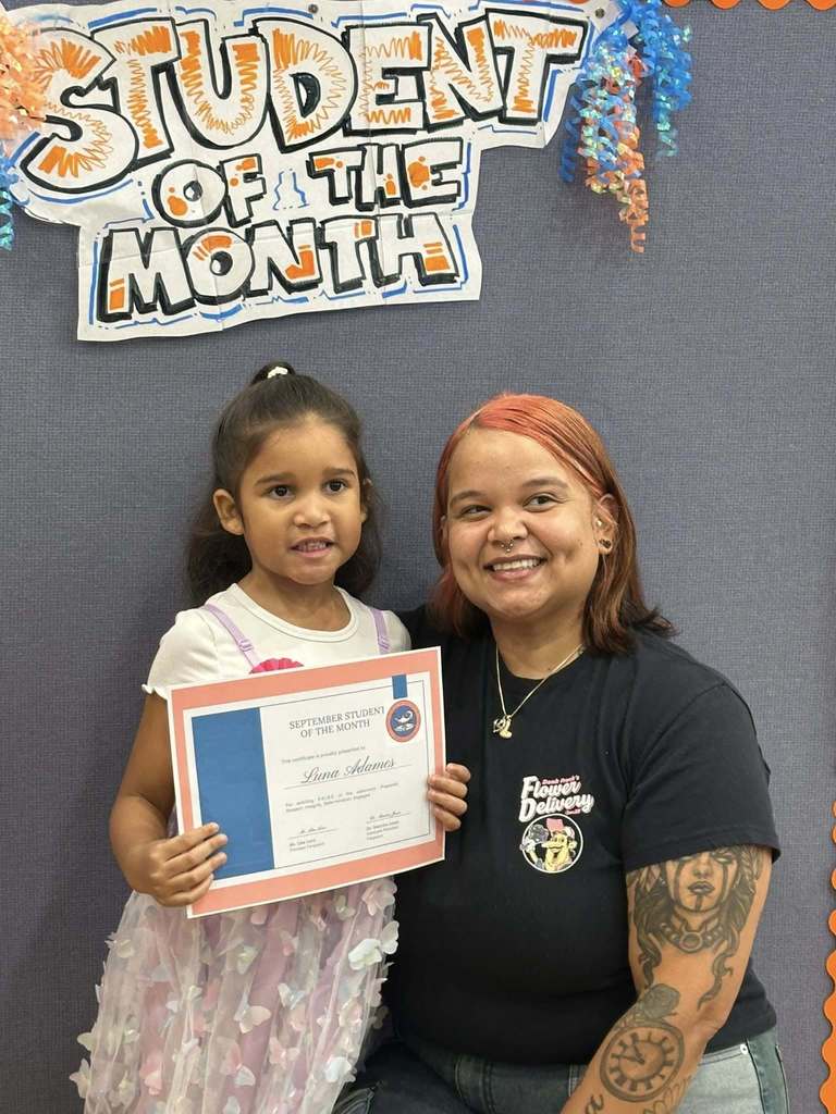 A student and an adult are smiling in front of a "Student of the Month" sign. The student is holding a certificate at Ferguson PreK-8's September Student of the Month Celebration.