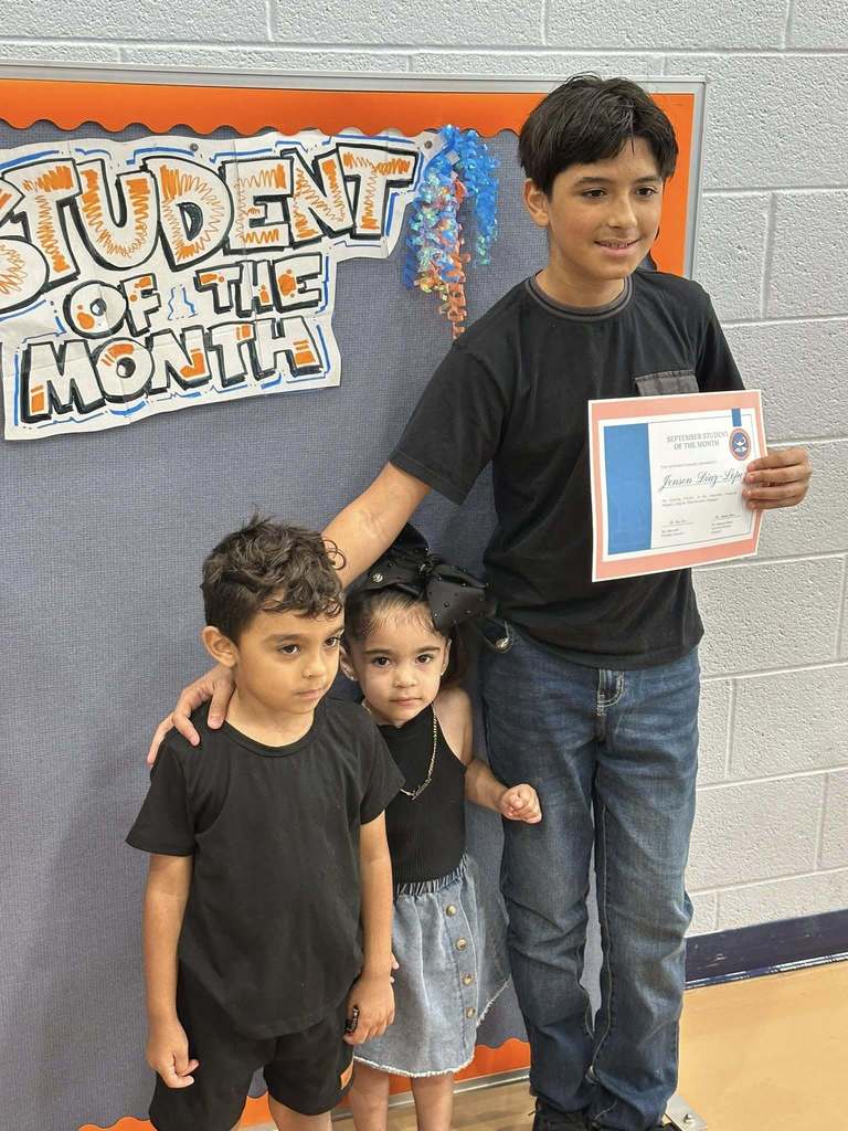 A student and two younger children are standing in front of a "Student of the Month" sign, holding a certificate at Ferguson PreK-8's September Student of the Month Celebration.