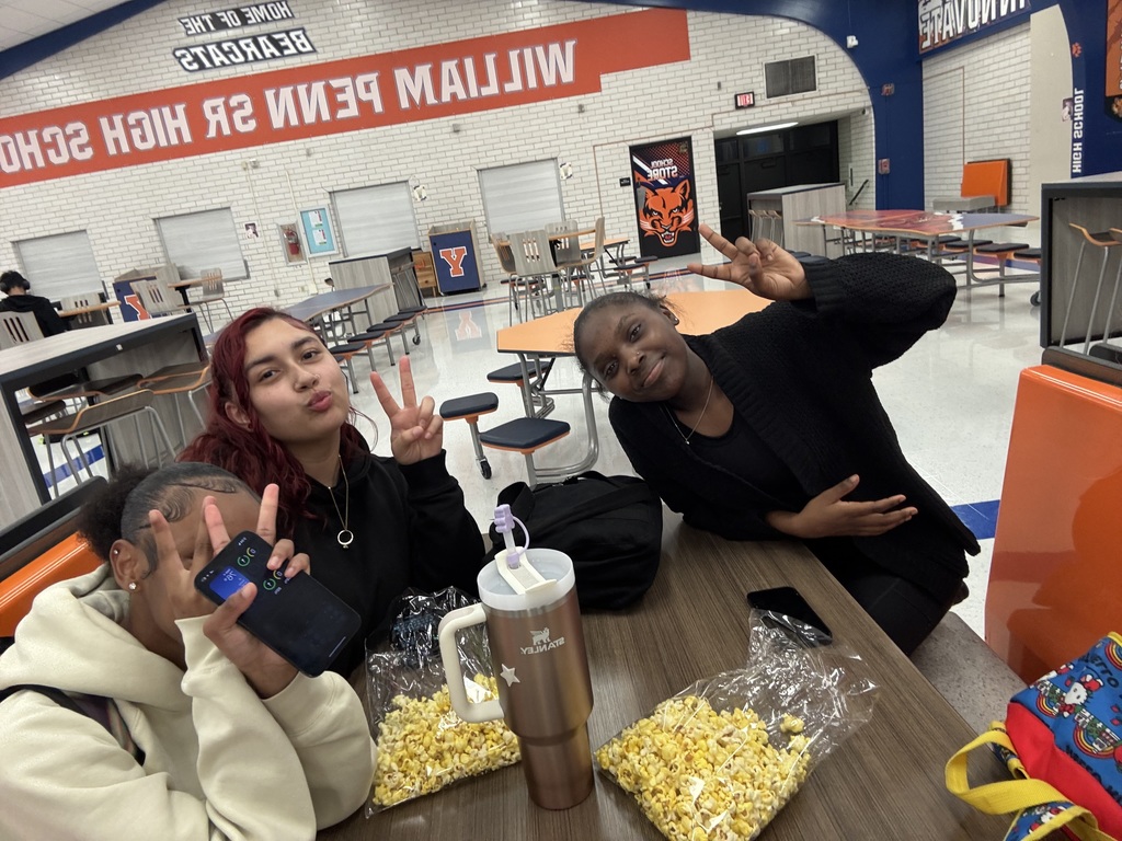 Three students smiling and flashing peace signs with bags of popcorn while sitting at a table in a school cafeteria. There is an orange sign nearby titled "WILLIAM PENN SR HIGH SCHOOL".