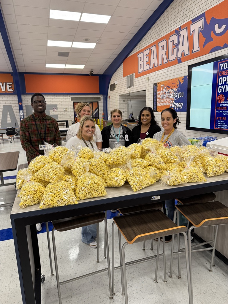 District staff standing behind a table piled with bags of popcorn in a school cafeteria. There is an orange sign nearby titled "BEARCAT."