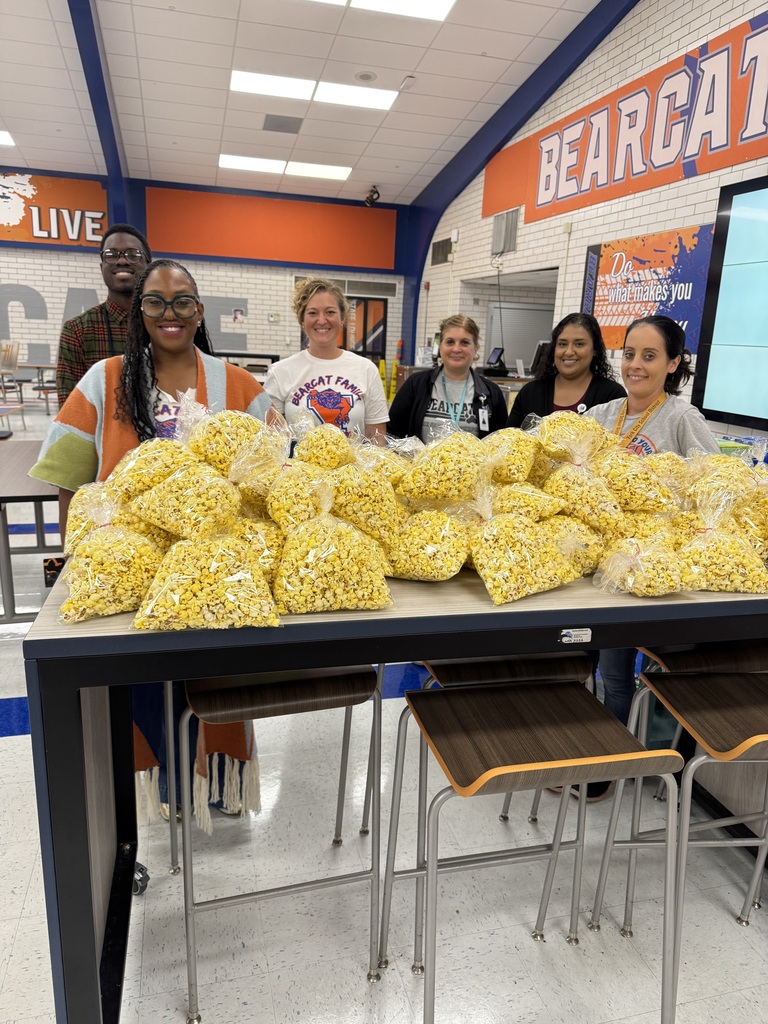 District staff standing behind a table piled with bags of popcorn in a school cafeteria. There is an orange sign nearby titled "BEARCAT."