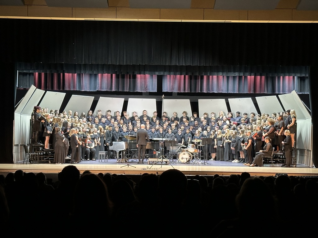 A large choir, accompanied by musicians and conductors, are performing on a stage in a well-lit auditorium.
