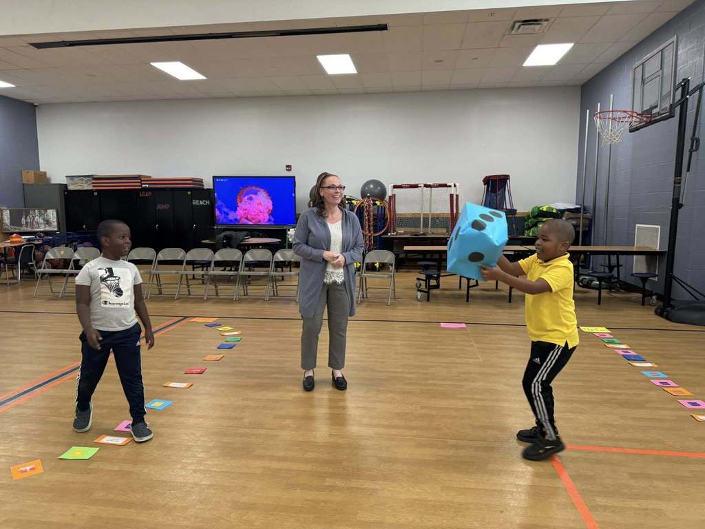 A district staff member and two young boys are playing in a school gym. One of the boys is catching a large blue die. Colorful cards can be seen nearby on the floor.
