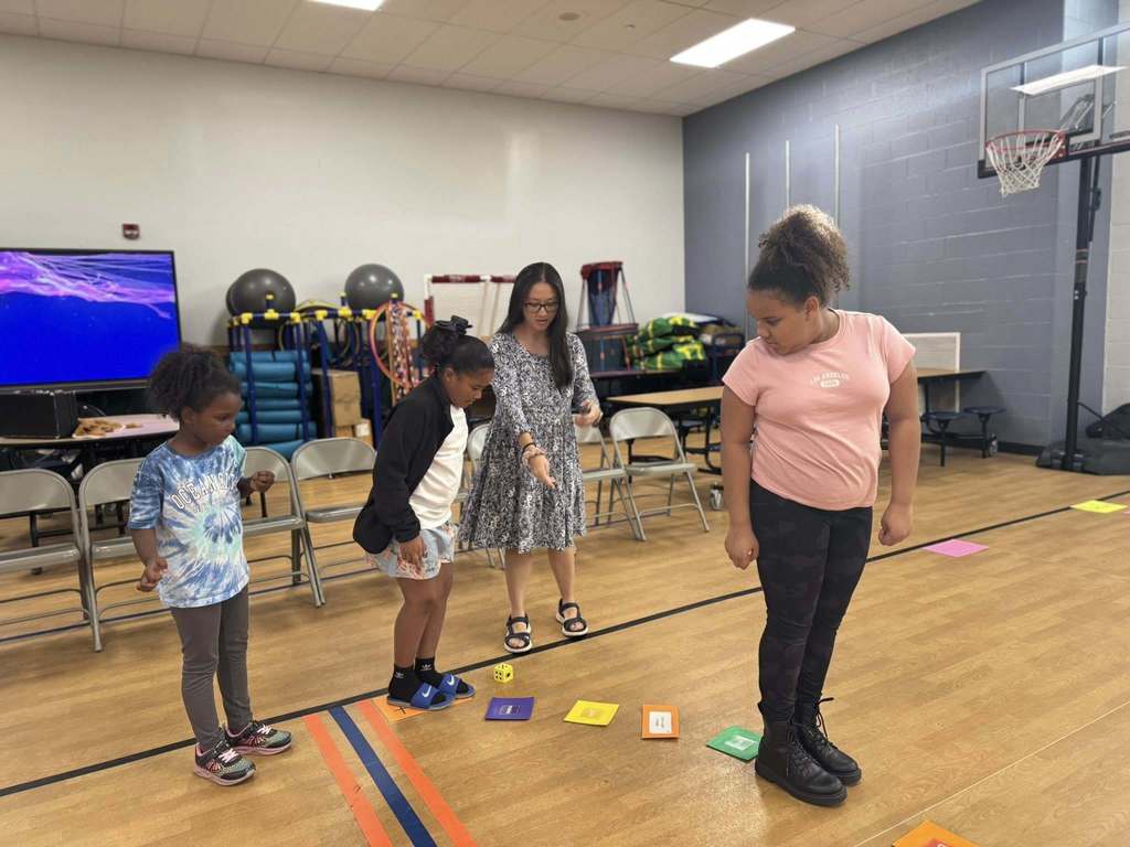 Three children and an adult are playing an educational game in a school gym. Colorful cards and dice are on the floor, creating a playful and engaging learning environment.