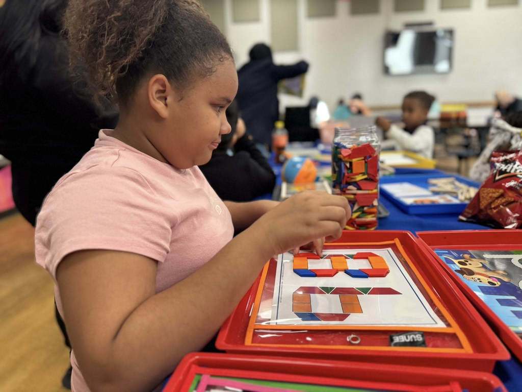 A young girl is focusing on assembling colorful geometric shapes in a school cafeteria. Other children and learning materials can be seen in the background. 