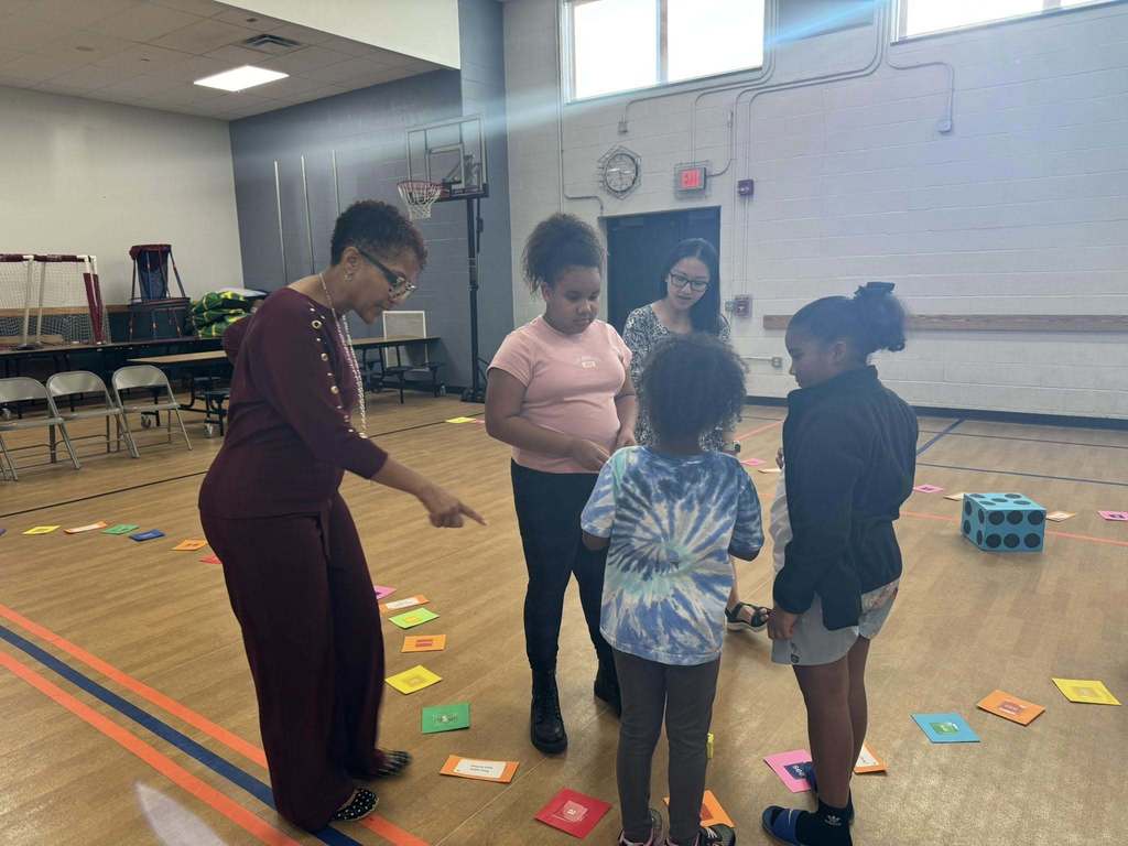 Jackson PreK-8 Principal, Ms. Smallwood and a district staff member are engaging with three young children in a school gym. Colorful cards and dice can be seen nearby on the floor. 